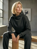 Model wearing Bou Clothing’s Black Marl luxury Deryn hoodie and matching joggers, seated on a wooden bench in a softly lit modern interior, showcasing a relaxed fit, premium fabric, and minimalist neutral style.