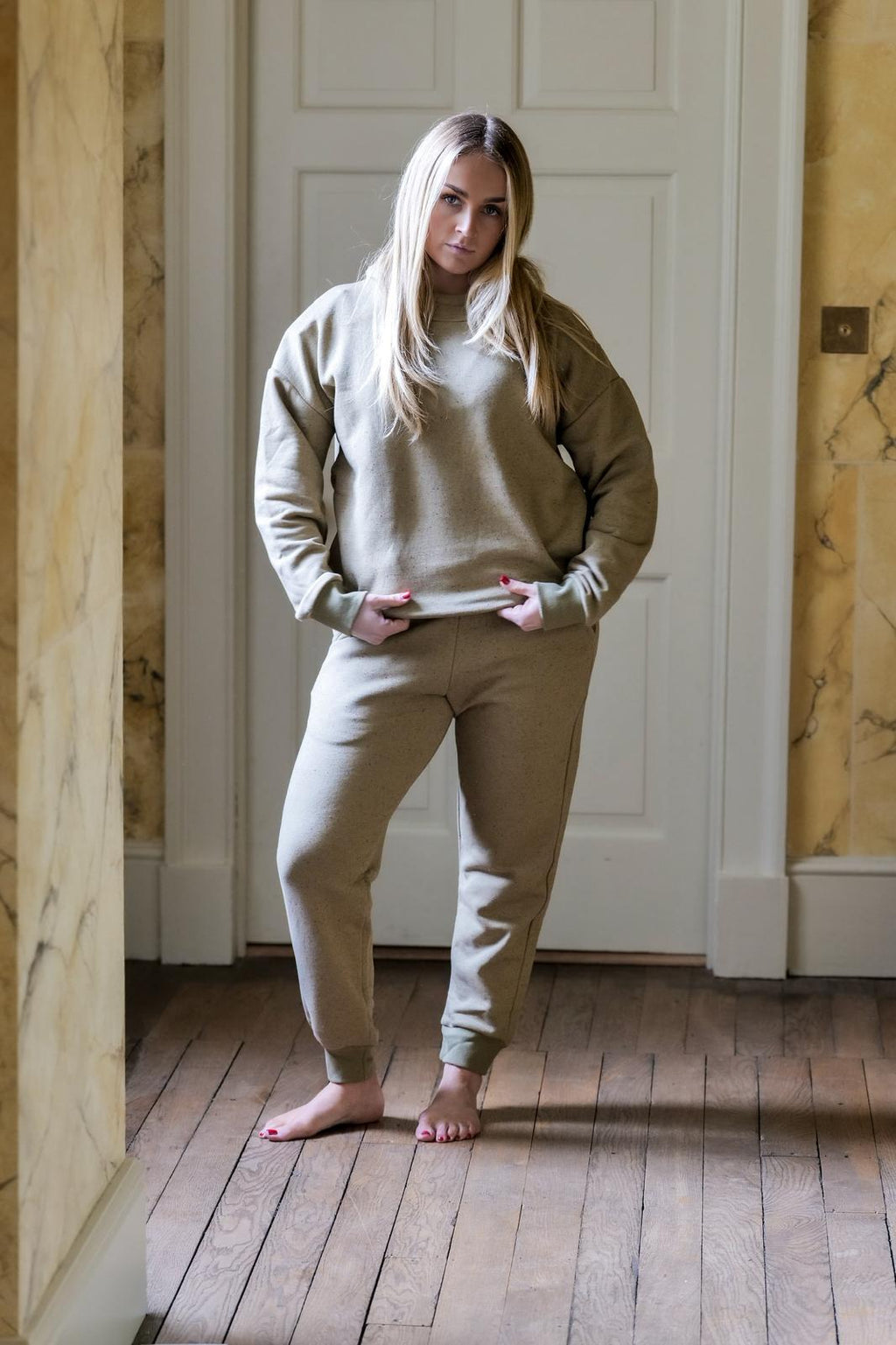 Woman wearing a beige tracksuit standing in a room with wooden floors and white walls.