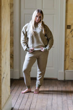Woman wearing a beige tracksuit standing in a room with wooden floors and white walls.