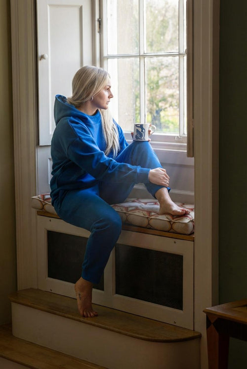 Woman sitting on a window seat with a cup, wearing a blue hoodie and pants.