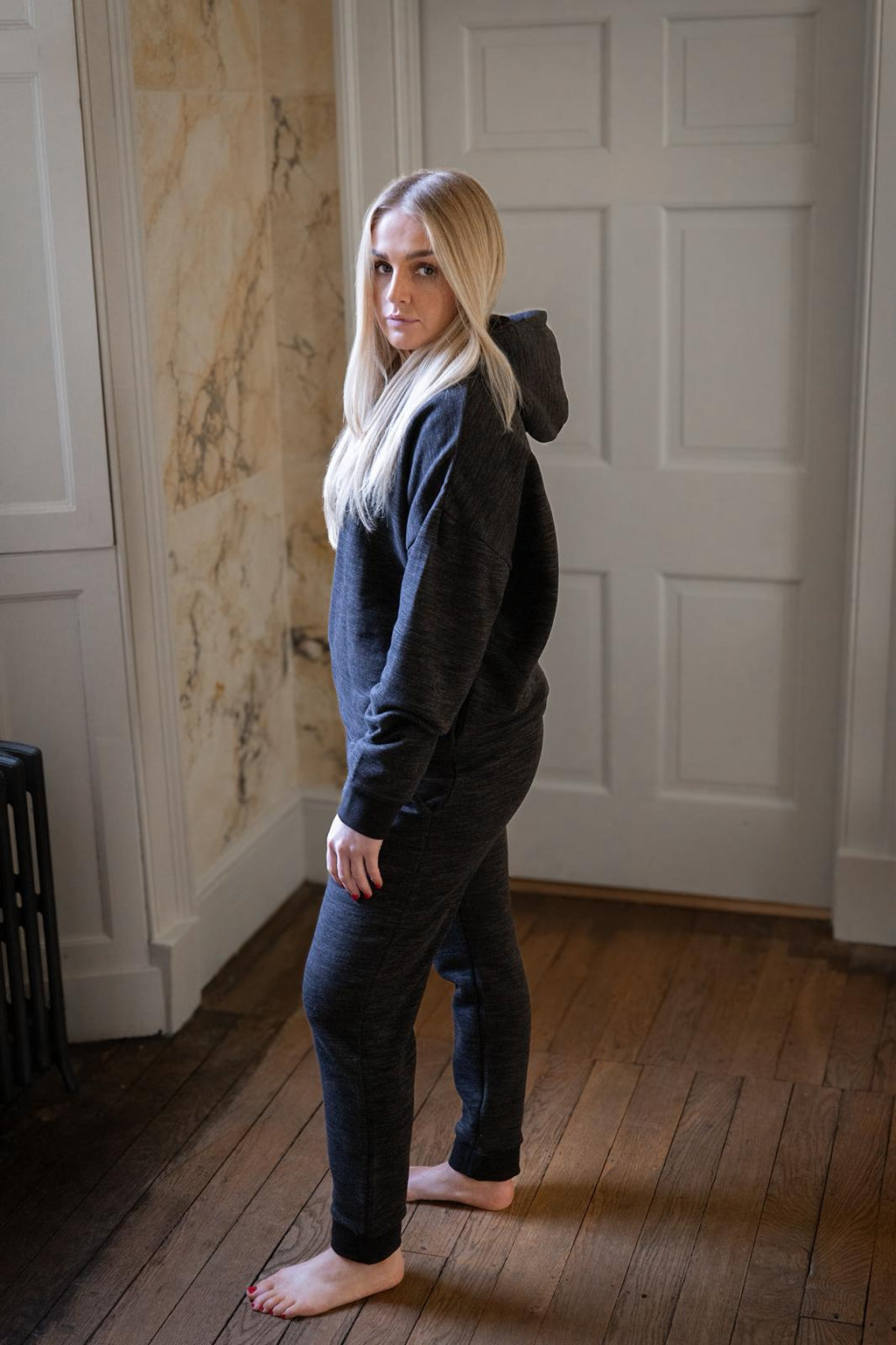 Woman wearing a dark hoodie and pants standing in a room with wooden flooring and white walls.
