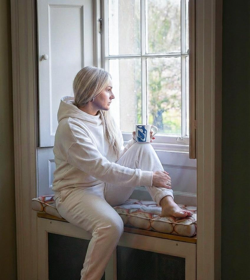 Woman sitting on a window seat holding a mug, wearing a white hoodie and pants.