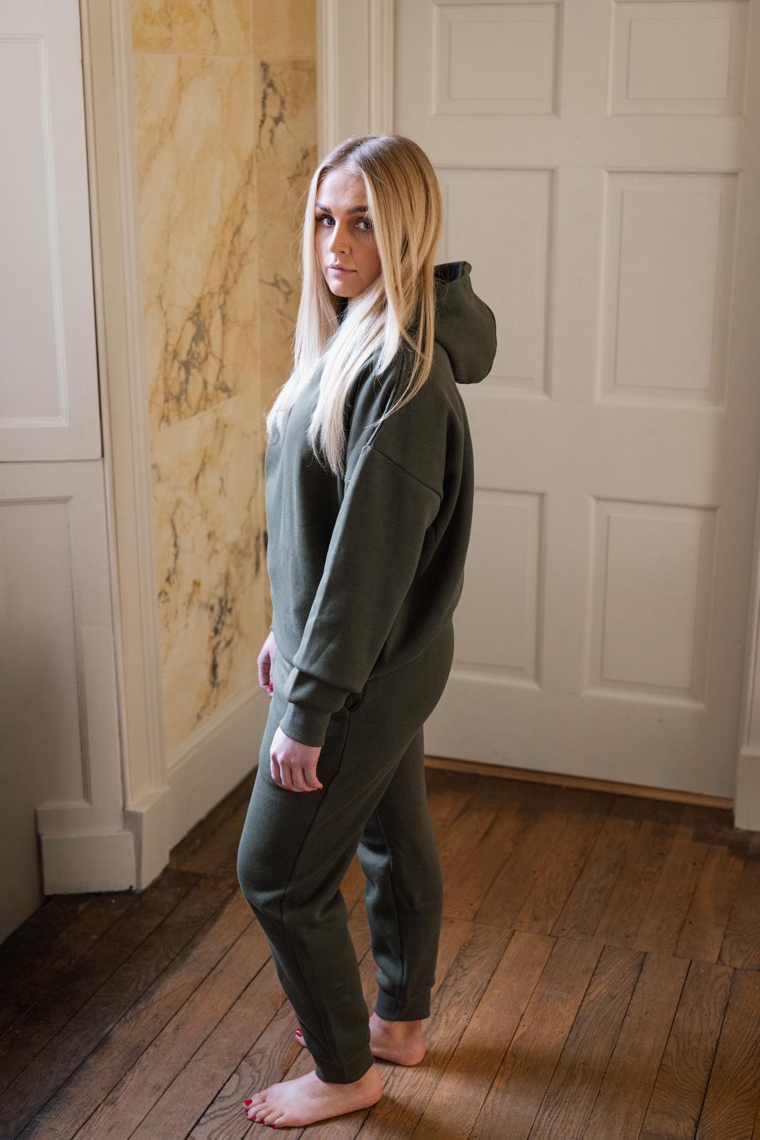 Woman wearing a green hoodie and pants standing in a room with wooden flooring and white walls.