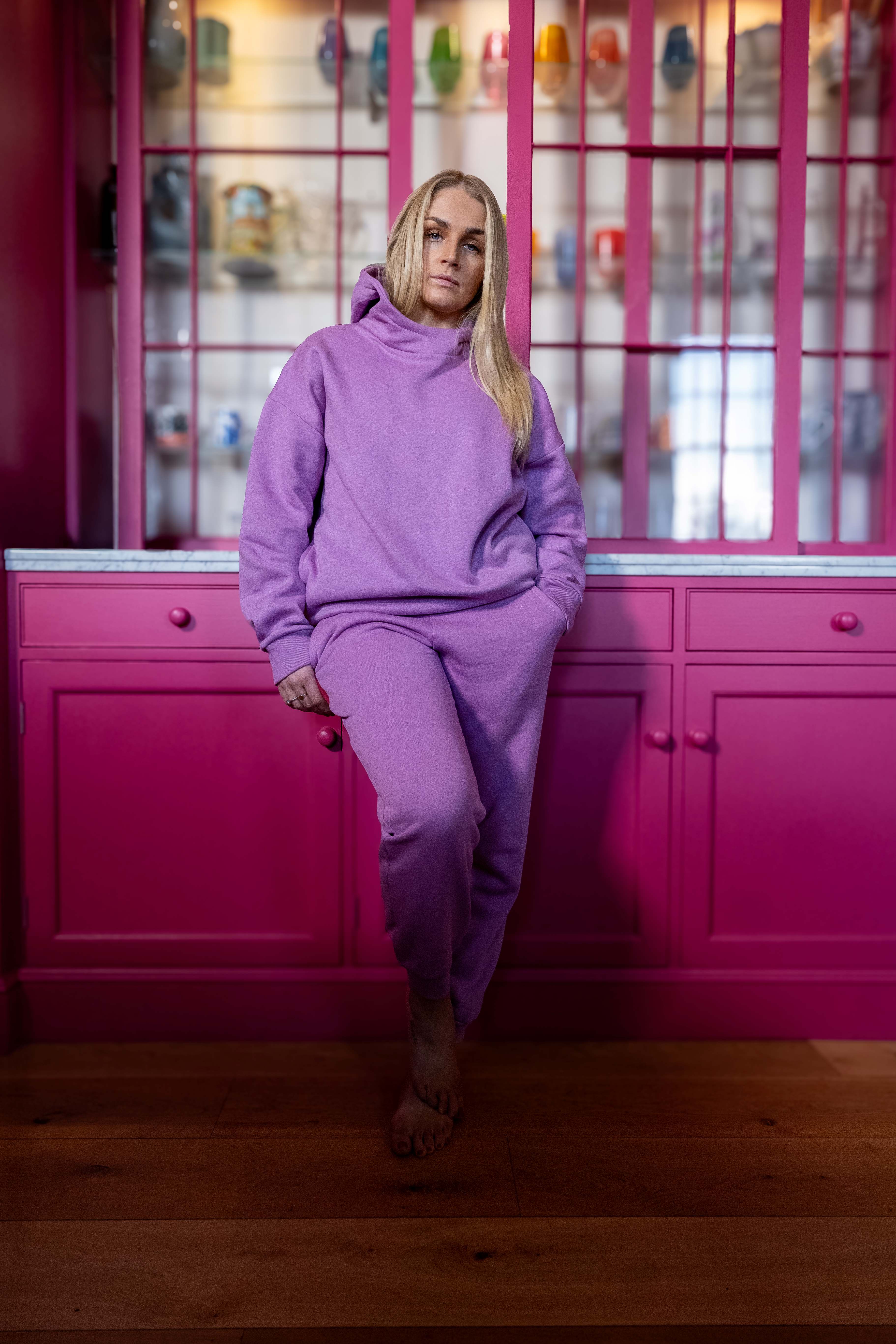 Person wearing a sorbet hoodiw and joggers outfit sitting against a pink kitchen cabinet with glass panels.