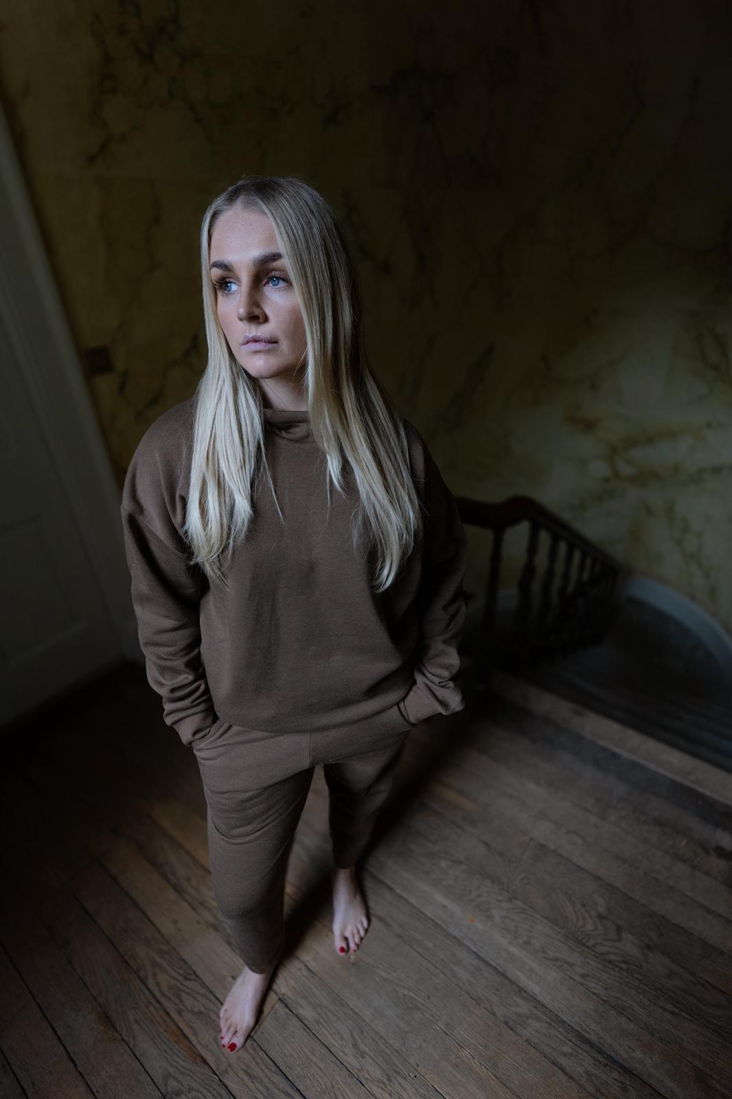 Woman in brown outfit standing in a dimly lit room with wooden floor and walls.