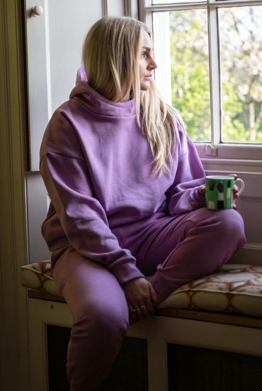 Person wearing a purple hoodie and pants sitting on a window sill holding a mug.