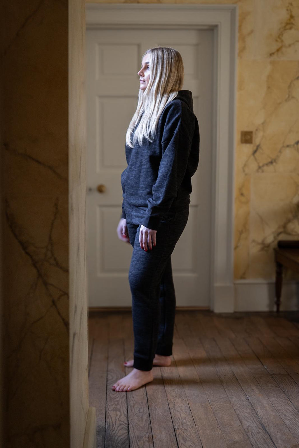 Person wearing a dark hoodie and pants standing in a room with marble walls.
